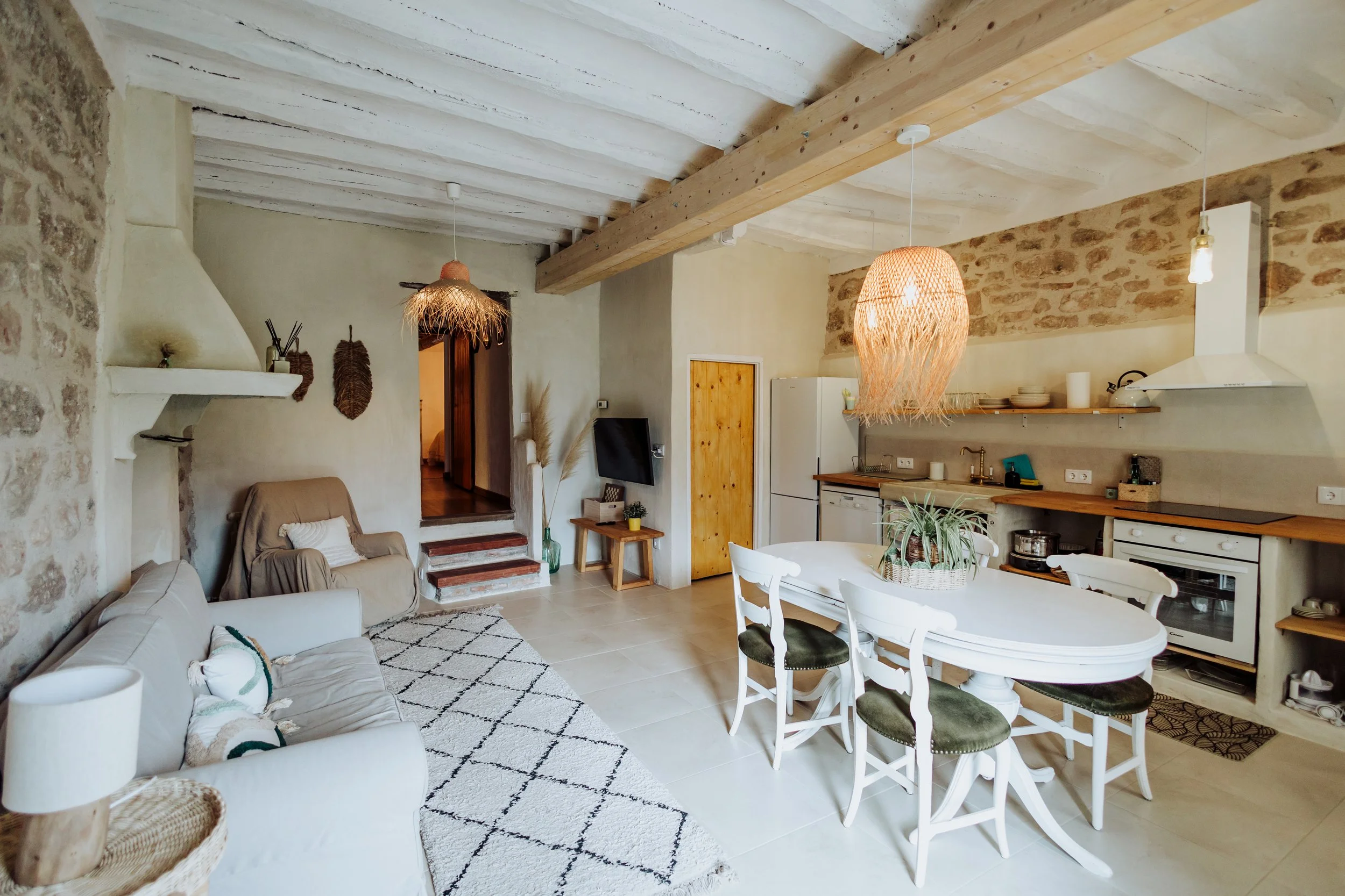 A cozy rustic living room with white walls, exposed wooden beams on the ceiling, and a mix of modern and vintage decor. It includes a beige sofa with pillows, a patterned rug, a small wooden side table with a plant, a wooden staircase, and hanging wicker lamps.