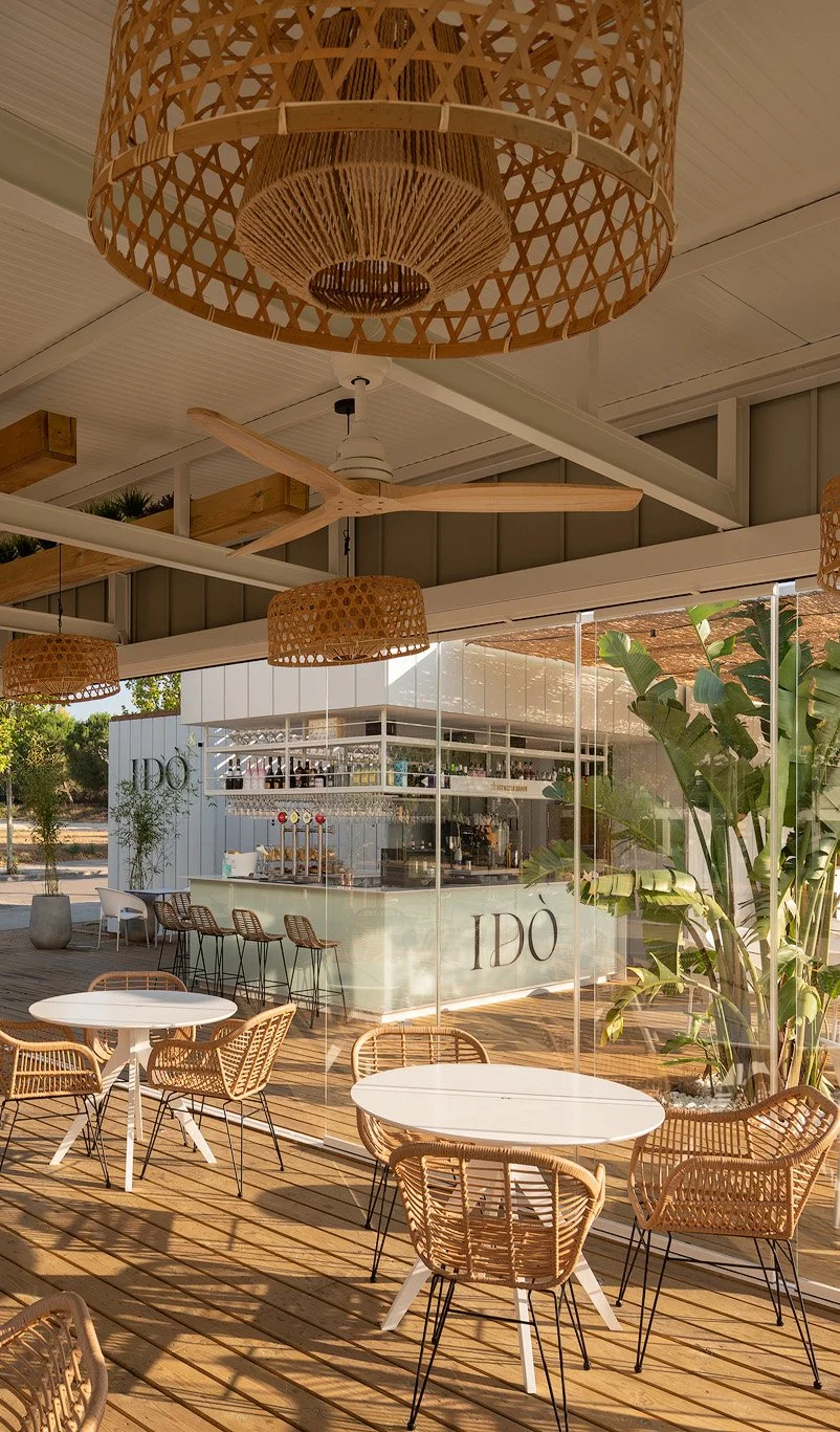 A stylish outdoor cafe with wicker chairs and white round tables. The area is shaded by wicker pendant lamps and has a view of a modern bar counter with the word 'IDO' on the side. Large green plants add to the relaxed atmosphere.