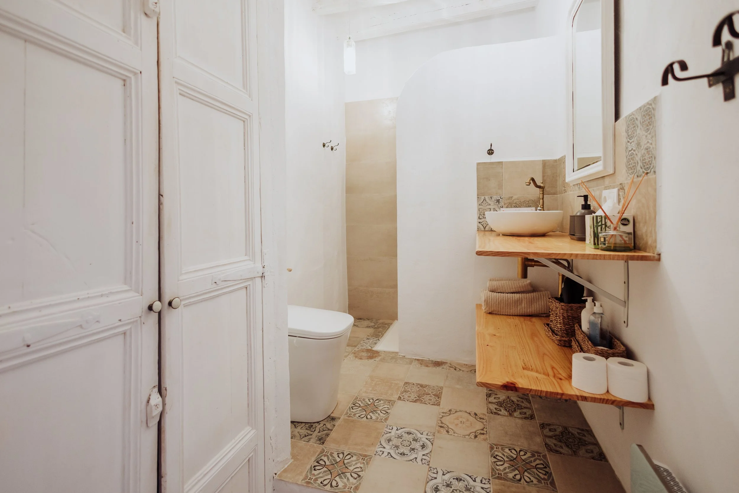 A cozy bathroom with a vintage white cabinet, a toilet, a walk-in shower with beige tiles, a wooden sink vanity with a round vessel sink, a mirror, and a shelf with toiletries, toilet paper, and decorative items.