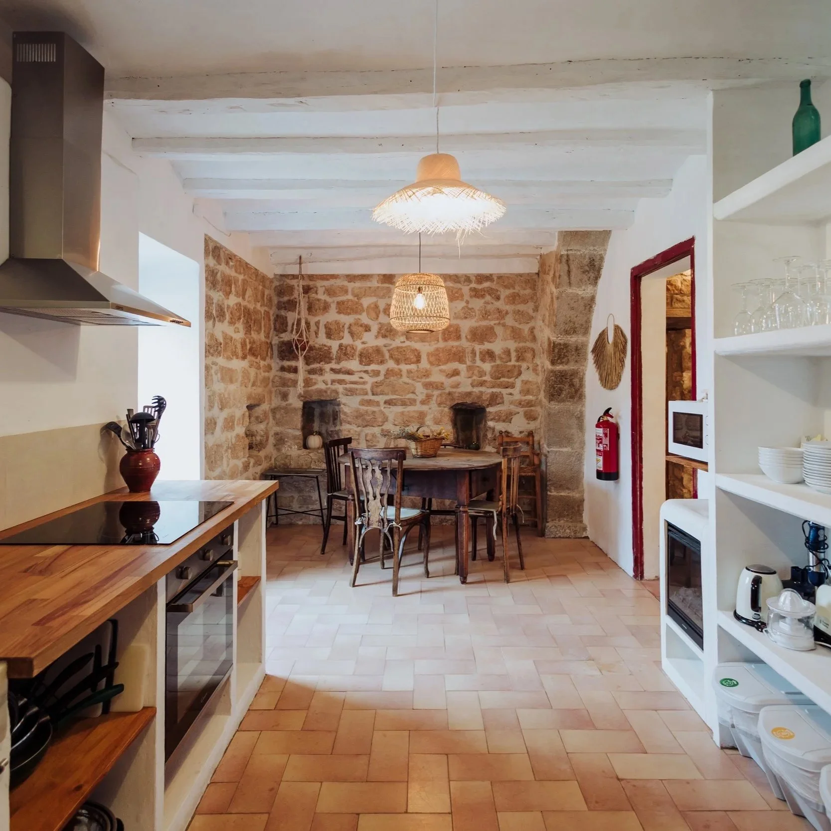 A cozy kitchen dining area with a stone accent wall, wooden dining table and chairs, hanging wicker light fixtures, white shelves with glassware and kitchen appliances, and a fire extinguisher on the wall.