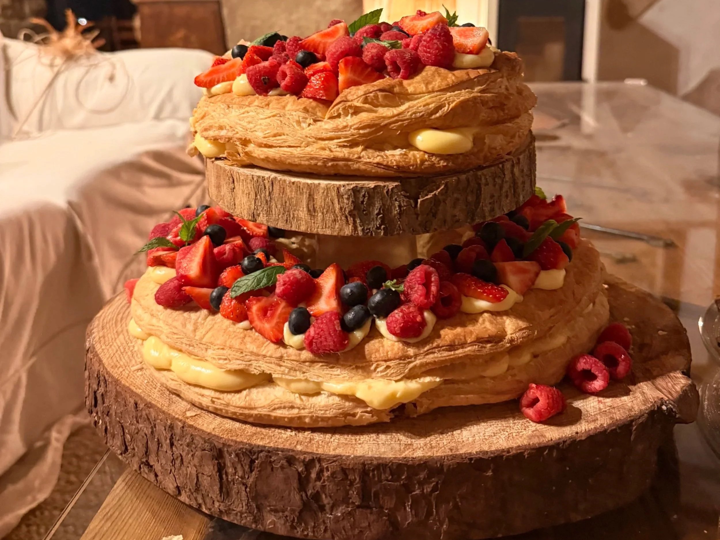 A three-tiered display of large homemade pavlova cakes topped with fresh strawberries, raspberries, blueberries, and mint leaves, set on a rustic wooden cake stand.