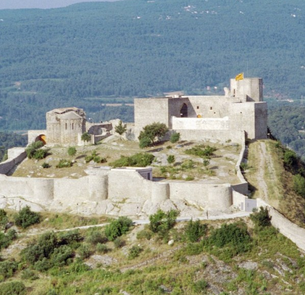A historic stone castle on a hill surrounded by greenery, with a flag on top of its highest tower.