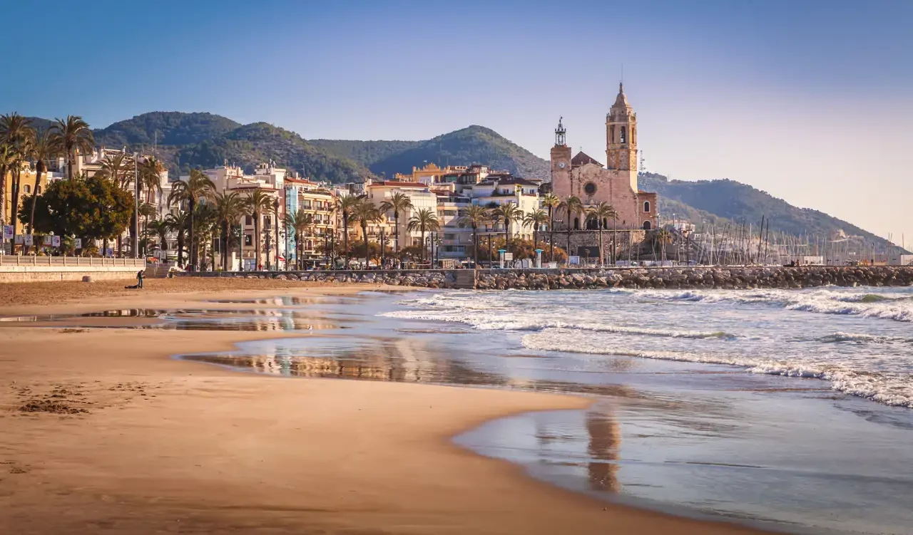 Beach with gentle waves, palm trees, colorful buildings, and a church tower in a coastal town with hills in the background.