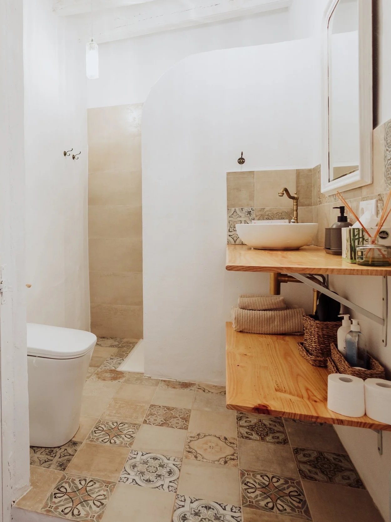 A cozy bathroom with a partial view of the toilet, a walk-in shower area with beige tile walls, a wooden vanity with a vessel sink, a large mirror, and decorative tiled flooring.