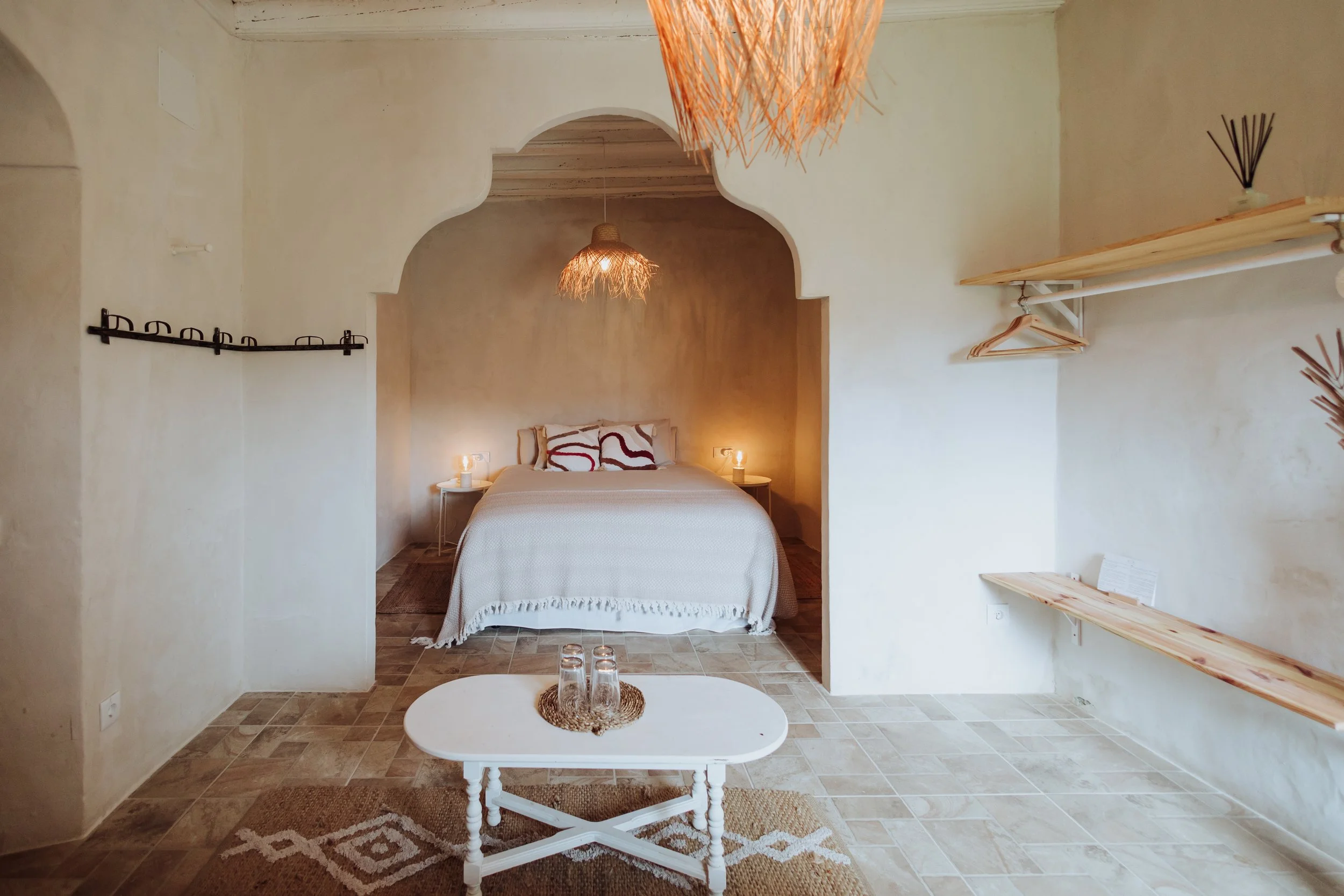 A bedroom with a bed, two nightstands with lamps, and decorative hanging lamps. The room has beige walls, wooden shelves, and a textured ceiling.