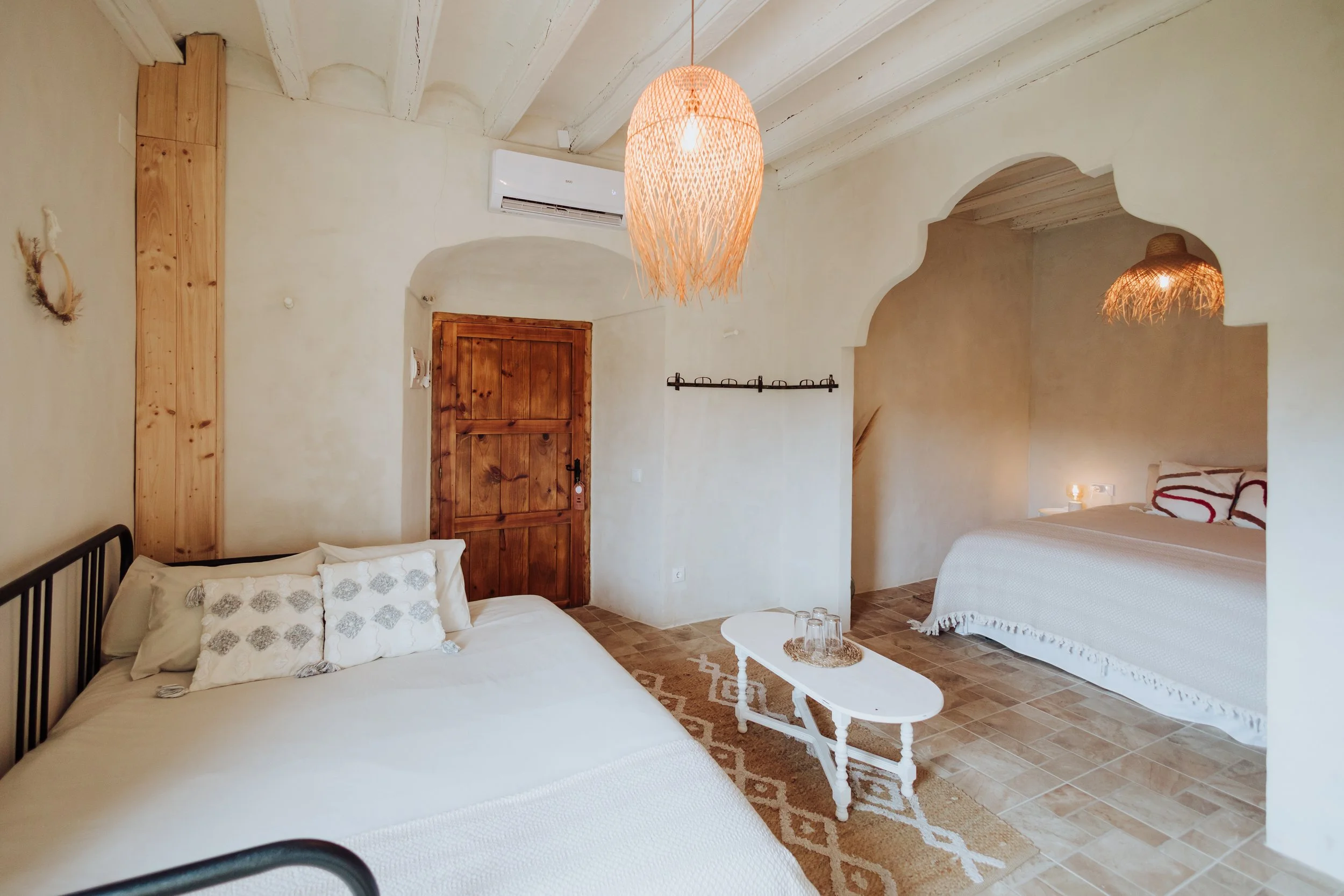 Interior of a cozy bedroom with white walls, a white beamed ceiling, and warm wooden accents. It features two beds with white bedding and decorative pillows, a small white coffee table, hanging woven pendant lights, and an air conditioning unit. The room has a rustic, Mediterranean style.