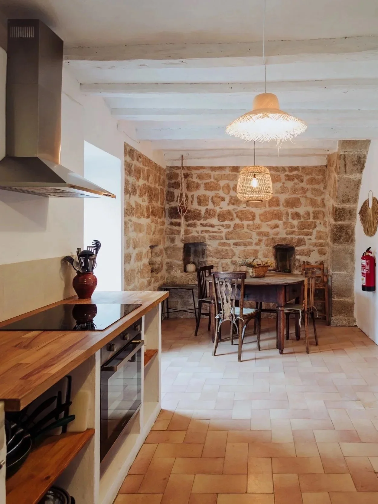 A rustic kitchen and dining area featuring a stone wall, wooden beams, and woven pendant lights. The kitchen has a wooden countertop with a red vase holding utensils, and a stove. The dining area has an oval wooden table with mismatched chairs and a corner fireplace.