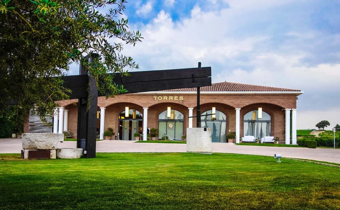 A building with a sign that says 'Torres', featuring large arched windows, surrounded by a well-manicured lawn and a large abstract black sculpture in the foreground.