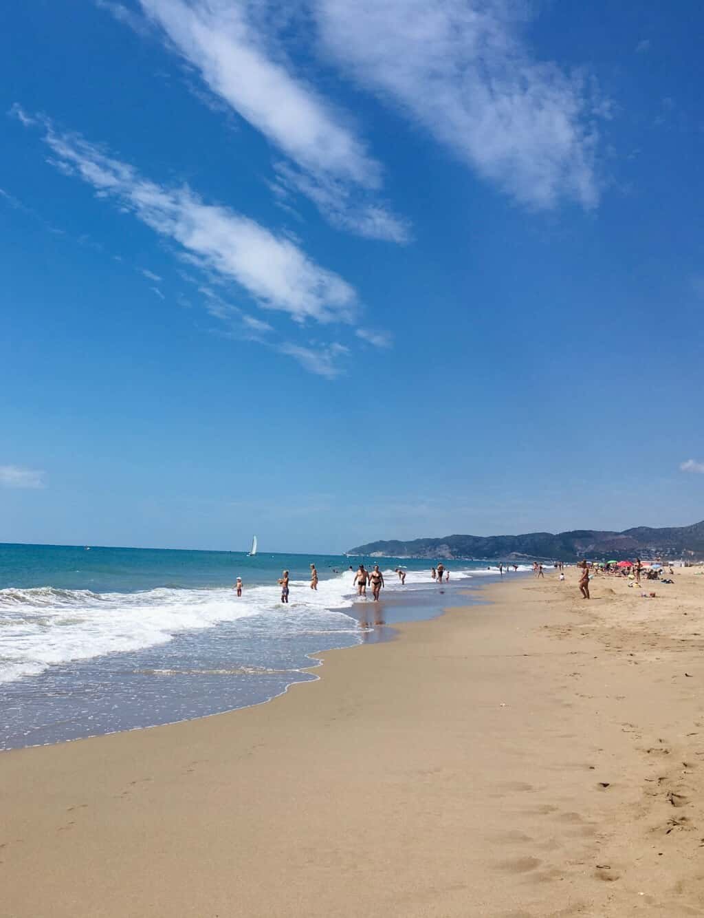 People swimming and walking on a sandy beach with blue sky, clouds, and mountains in the background. Sailboat visible on the water.