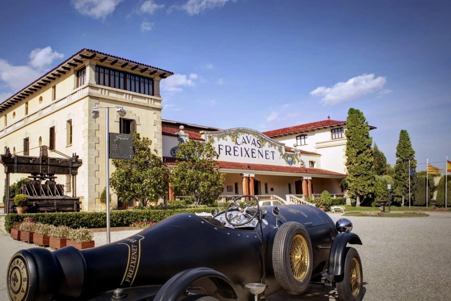A vintage race car parked in front of the Freixenet winery building with a sign reading 'Cavas Freixenet' and a scenic backdrop of trees and flags under a blue sky.