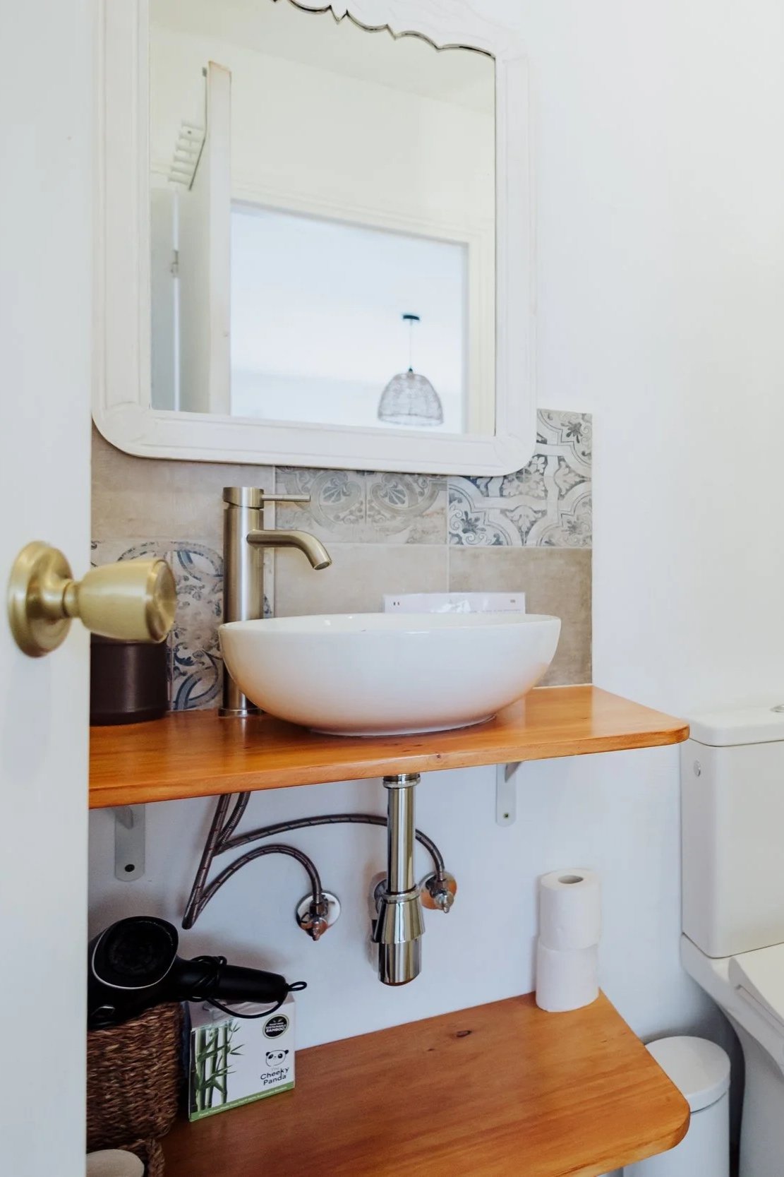 A small bathroom with a round vessel sink on a wooden countertop, a mirror above, and a faucet. There is a toilet to the right, and various items beneath the countertop, including a roll of toilet paper, a black basket, and a green box. The wall behind the sink has a decorative tile strip.