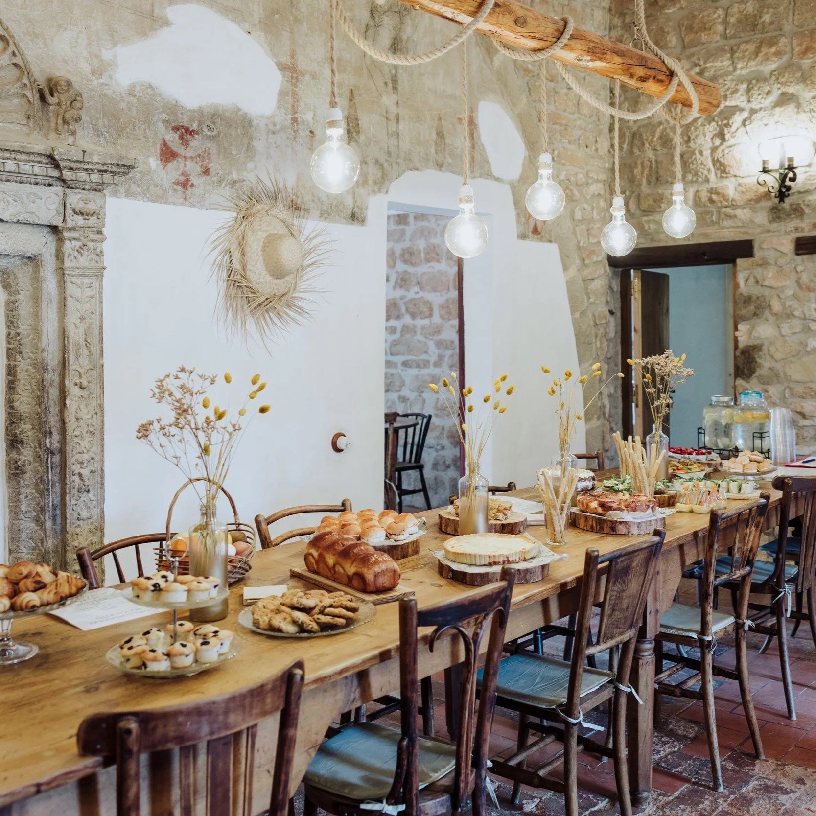 Rustic dining room with a long wooden table set with various baked goods and desserts, including muffins, bread, and cookies. The background features a stone wall with warm lighting from hanging light fixtures and a wall sconce, along with decorated dried flowers and a straw hat on the wall.