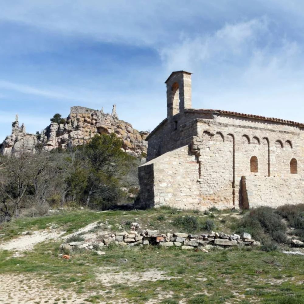 An old stone church with a bell tower on a hill, surrounded by sparse desert vegetation, with rocky cliffs in the background under a partly cloudy sky.