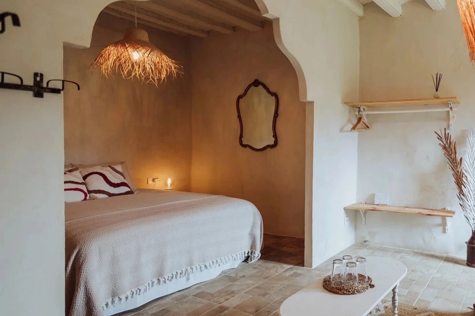 Cozy bedroom with rustic decor, featuring a bed with patterned pillows, a warm pendant light, wall mirror, and shelves with hangers on beige textured walls.