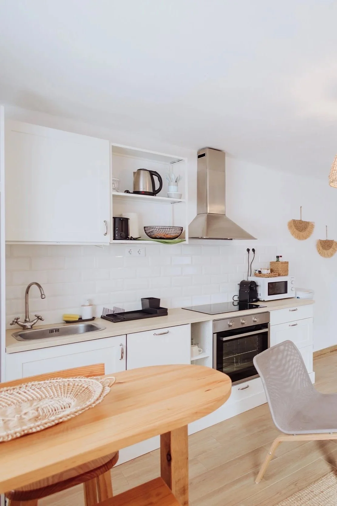 A modern, minimalist kitchen with white cabinets, a stainless steel range hood, a microwave, a coffee maker, and a wooden dining table with a woven placemat and a white chair.