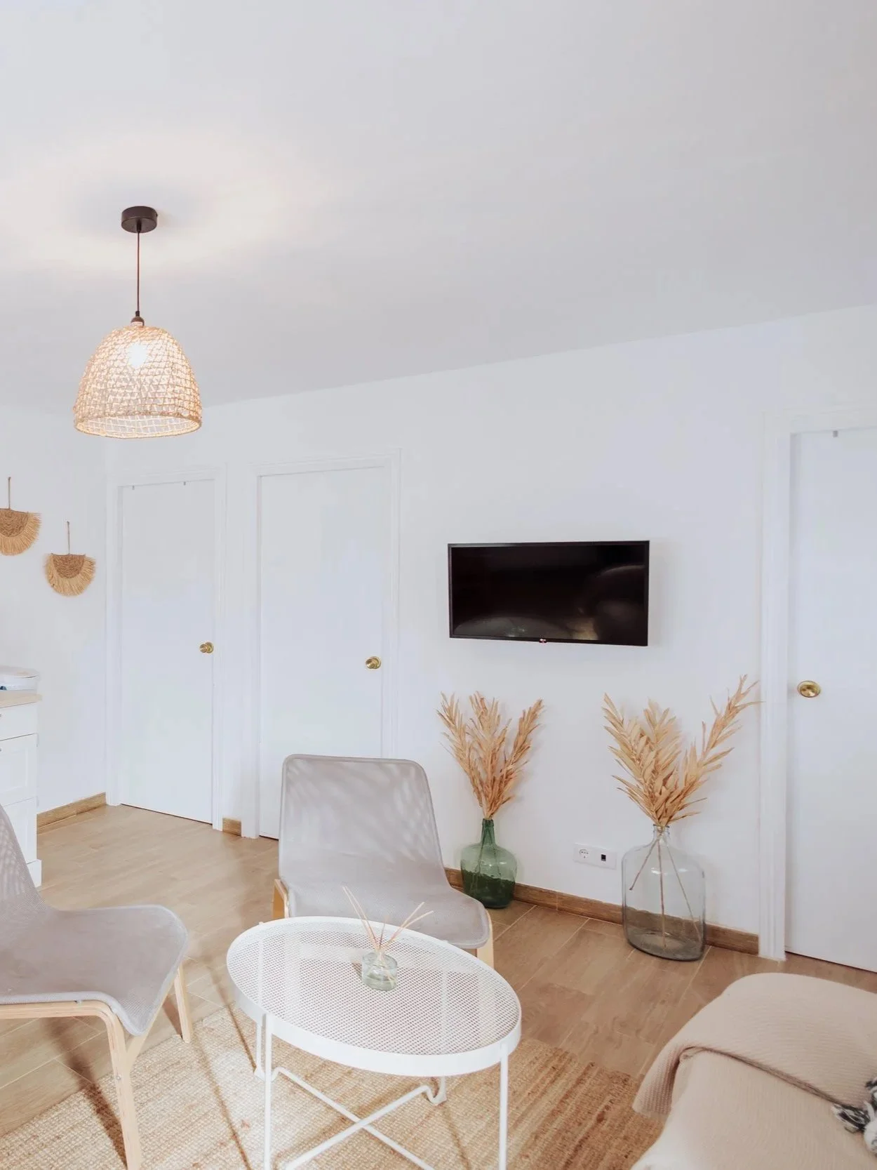 Bright living room with white walls, a wall-mounted TV, dried pampas grass in vases, beige chairs, a white round coffee table, and a woven pendant lamp hanging from the ceiling.