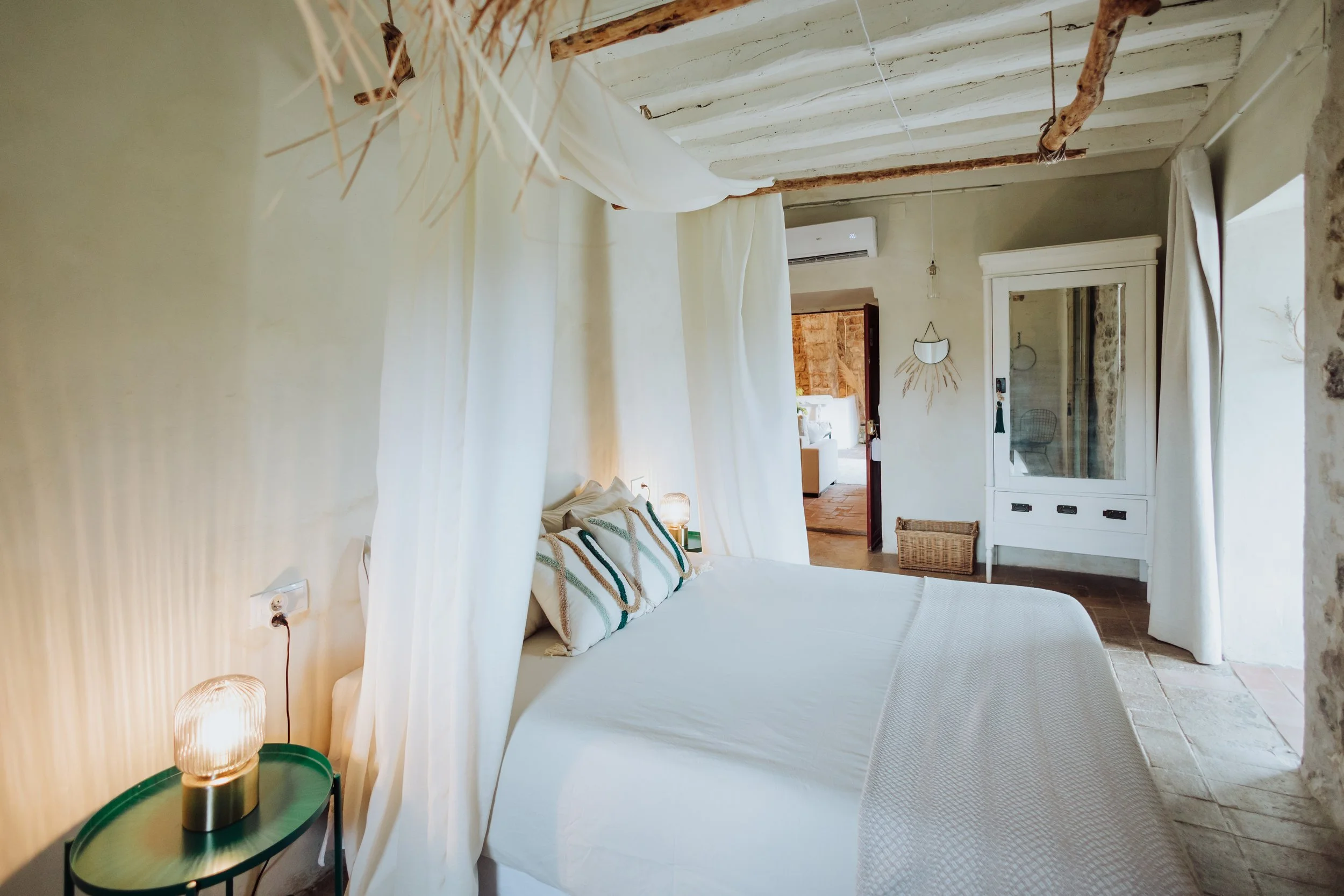 A bedroom with white curtains around a bed, pillows, and a bedside table with a lamp. The room has exposed wooden beams and a white wall with an air conditioning unit. There is a mirror and a door leading to another room.