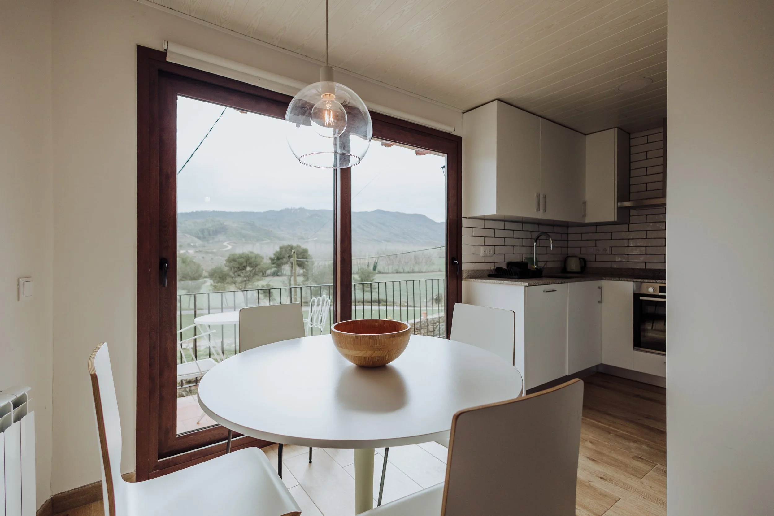 A dining area with a round white table surrounded by white chairs, positioned near large glass sliding doors with a mountain view outside. There is a wooden bowl on the table. The kitchen in the background has white cabinets, a black faucet, and a brick-style backsplash.