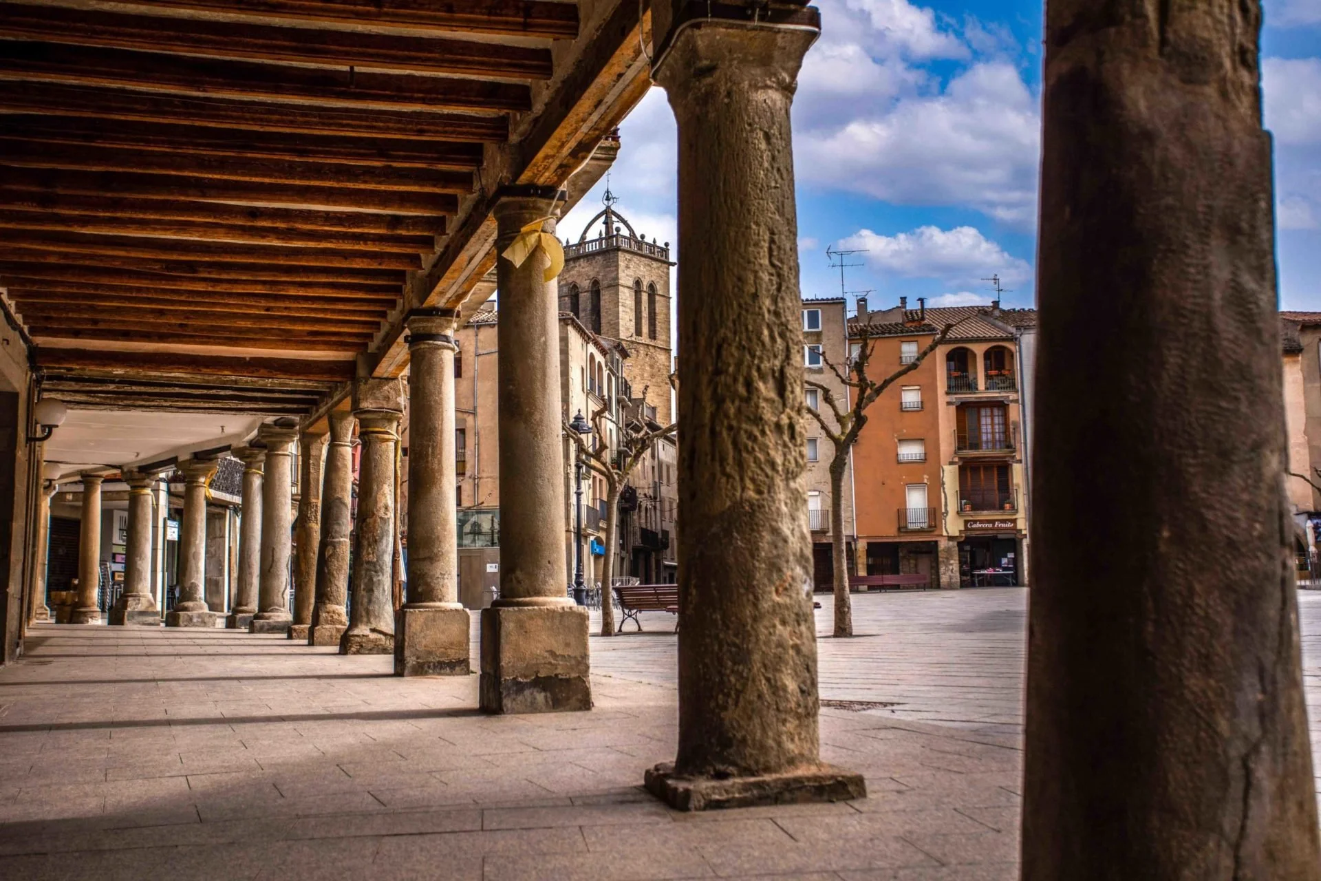View of an outdoor historic European square with stone columns supporting a covered walkway, surrounding buildings with balconies and antennas, some trees, a cloudy blue sky, and benches.