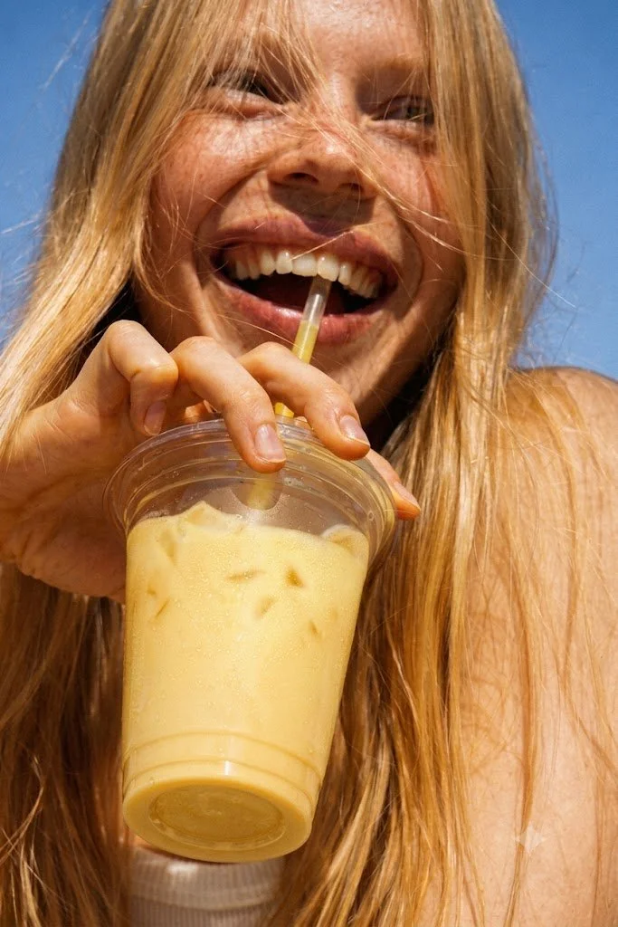 A woman with long blonde hair is enjoying an iced yellow safra latte with a straw.