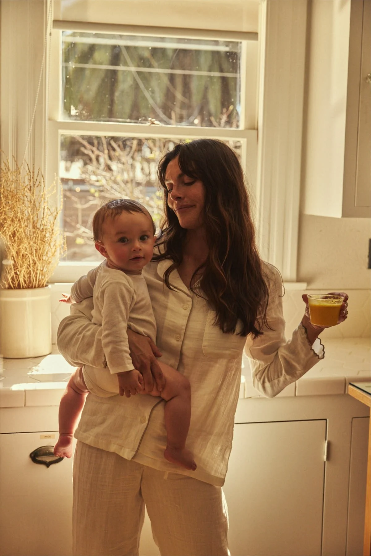A woman with long dark hair holding a young child in a beige outfit in a kitchen with a window, a potted dried plant, and a mug with a yellow safra latte drink.