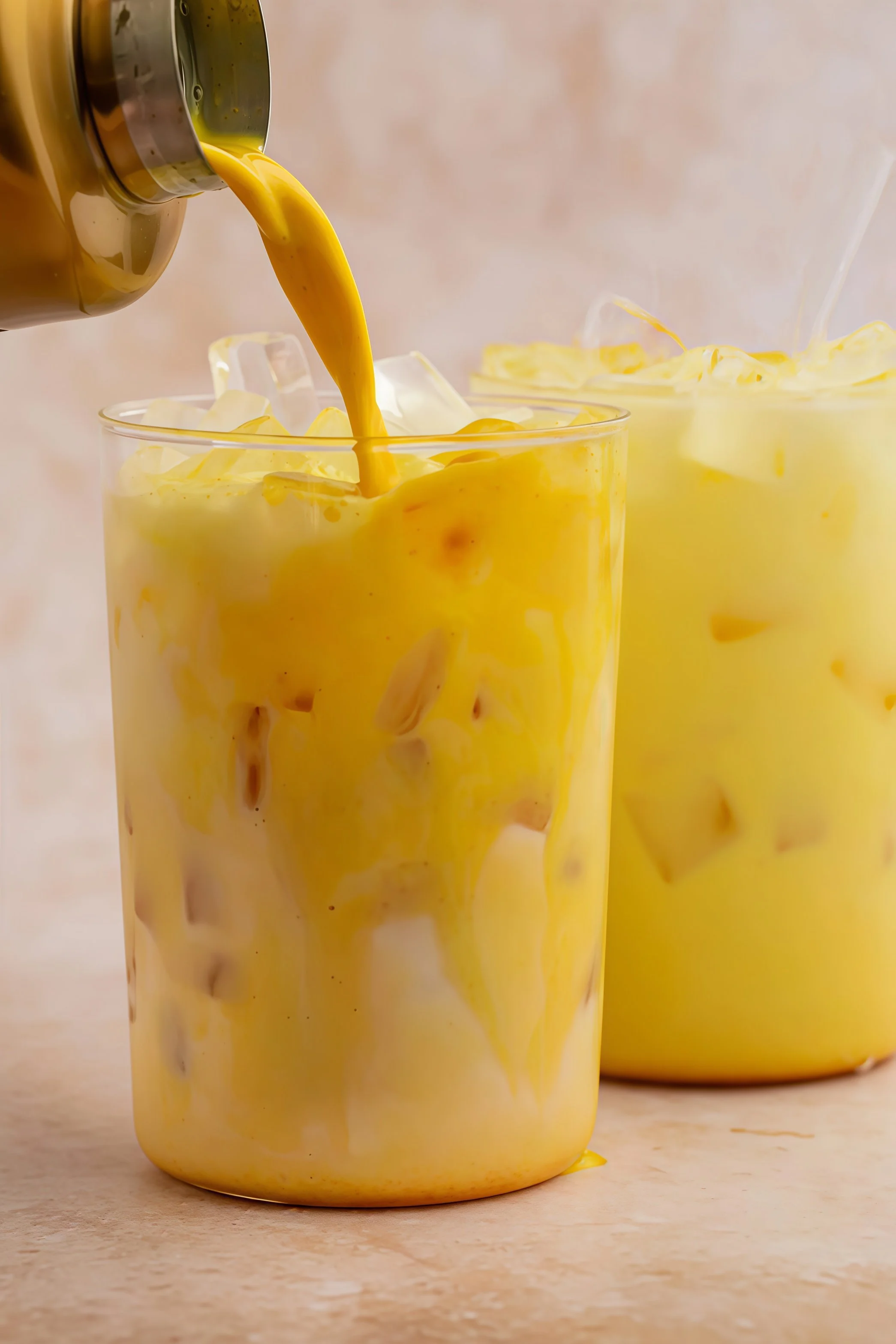 Yellow safra latte being poured into a glass with ice cubes, with a second glass in the background.