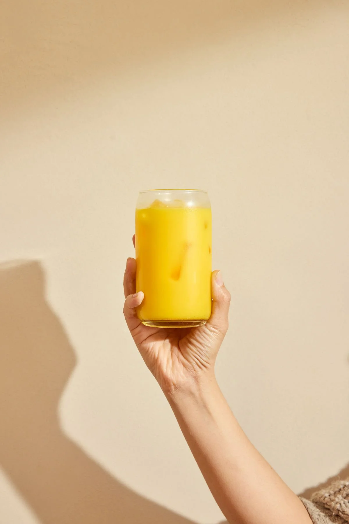 A person holding a glass of yellow safra latte with ice and pieces of fruit against a plain beige wall.