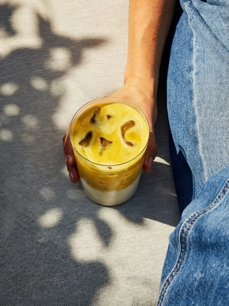 A person is sitting on a light-colored surface, holding a glass of iced safra latte with cream and ice cubes, outdoors in sunlight.