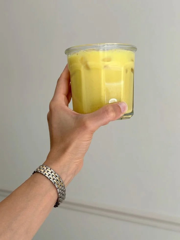 A hand holding a glass container filled with yellow safra latte, against a plain wall background.