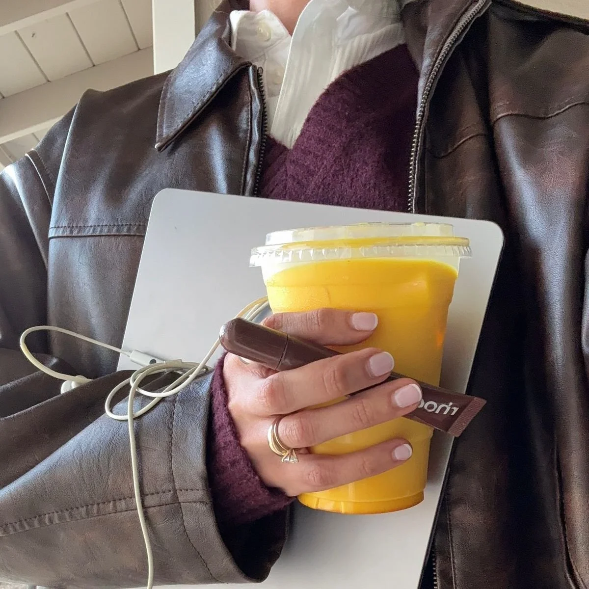 Person wearing a brown leather jacket, white shirt, and purple sweater holding a yellow safra latte, a brown rhode lipstick, and some electronic devices with earphones.