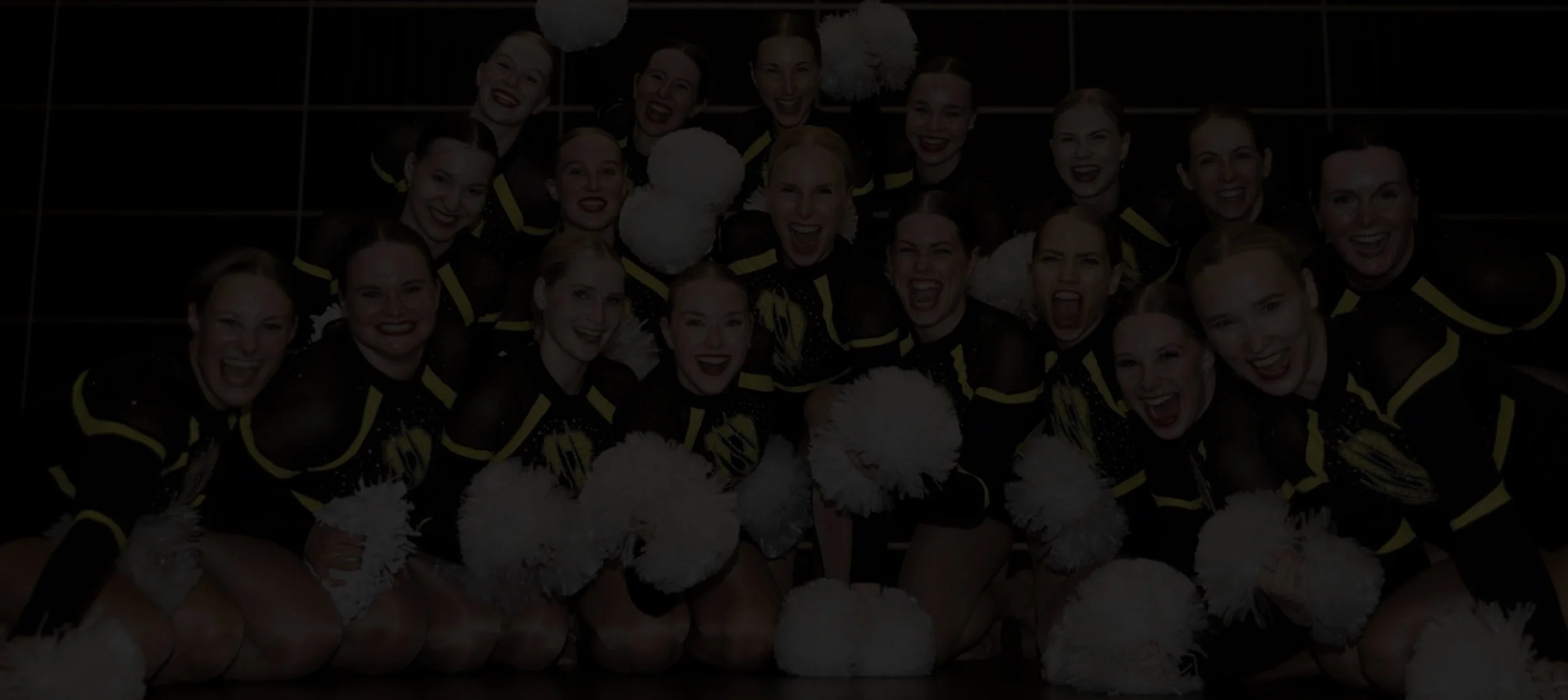Ryhmä cheerleadereita istuu lattialla, hymyilee ja pitää valkoisia viuhkoja, taustalla pimeä seinä.