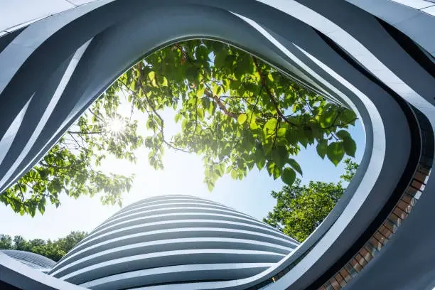 Looking up through a modern building's open roof, with sunlight and green trees visible.