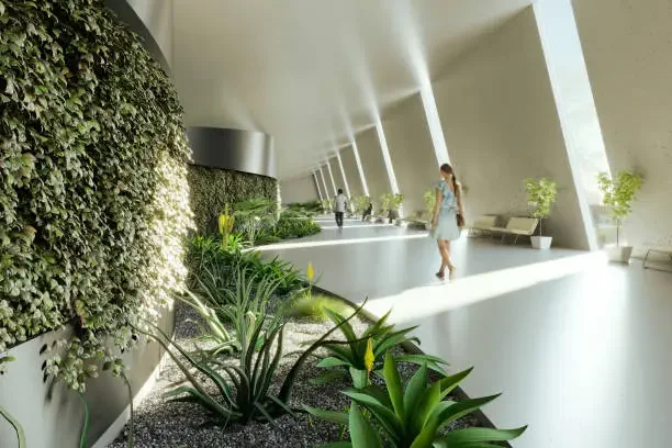 Modern indoor space with large windows, sunlight, greenery, benches, and a few people walking.