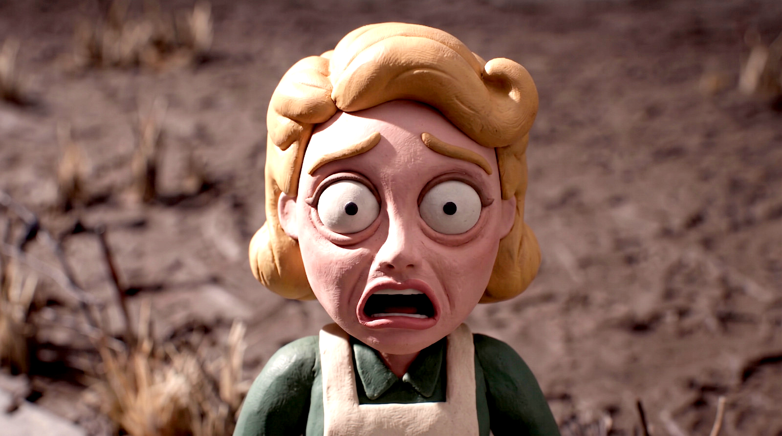 A claymation character with wide eyes and a shocked expression, with blonde hair and wearing a green shirt with a beige apron, standing outdoors in dirt.