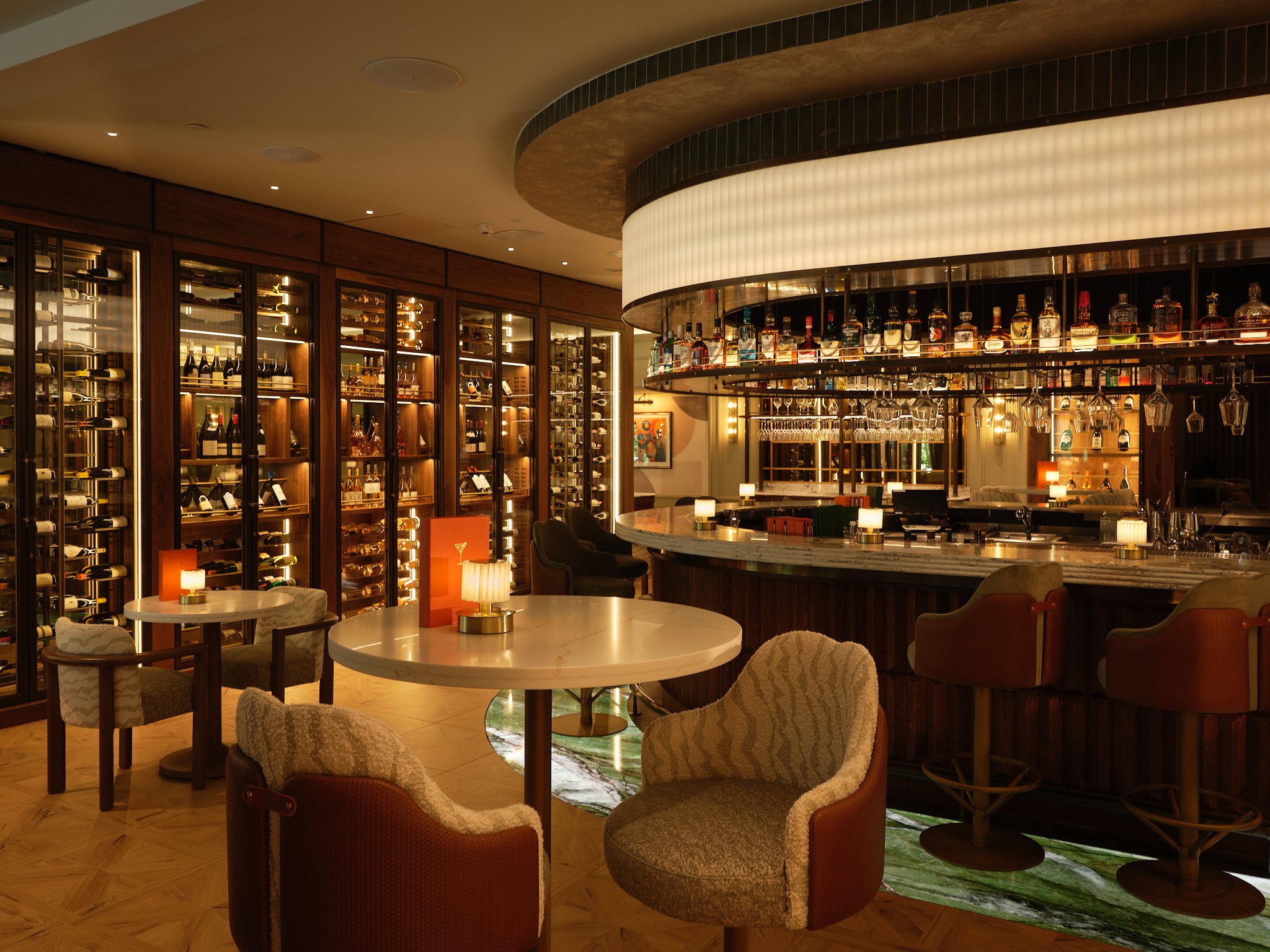 Inside a modern bar with a curved wine display shelf filled with various liquor bottles, glassware, round tables, and upholstered chairs.