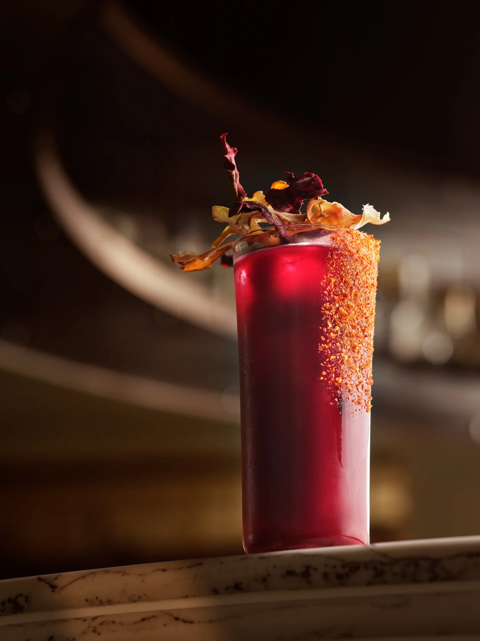 A close-up of a tall, vibrant red cocktail garnished with dried fruit and flower petals, in a cylindrical glass on a marble surface.