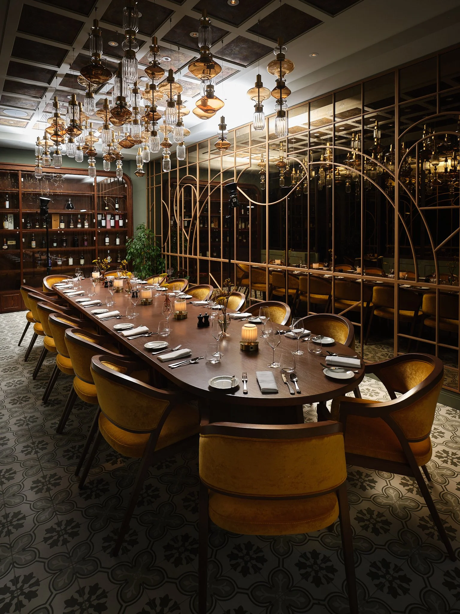 Elegant dining room with a large wooden table set with plates, glasses, napkins, and candles. Yellow velvet chairs surround the table. Overhead, a modern chandelier with multiple glass and bronze fixtures hangs. The background features wooden shelves with bottles and a decorative metal partition, with mirrors reflecting the room's decor.