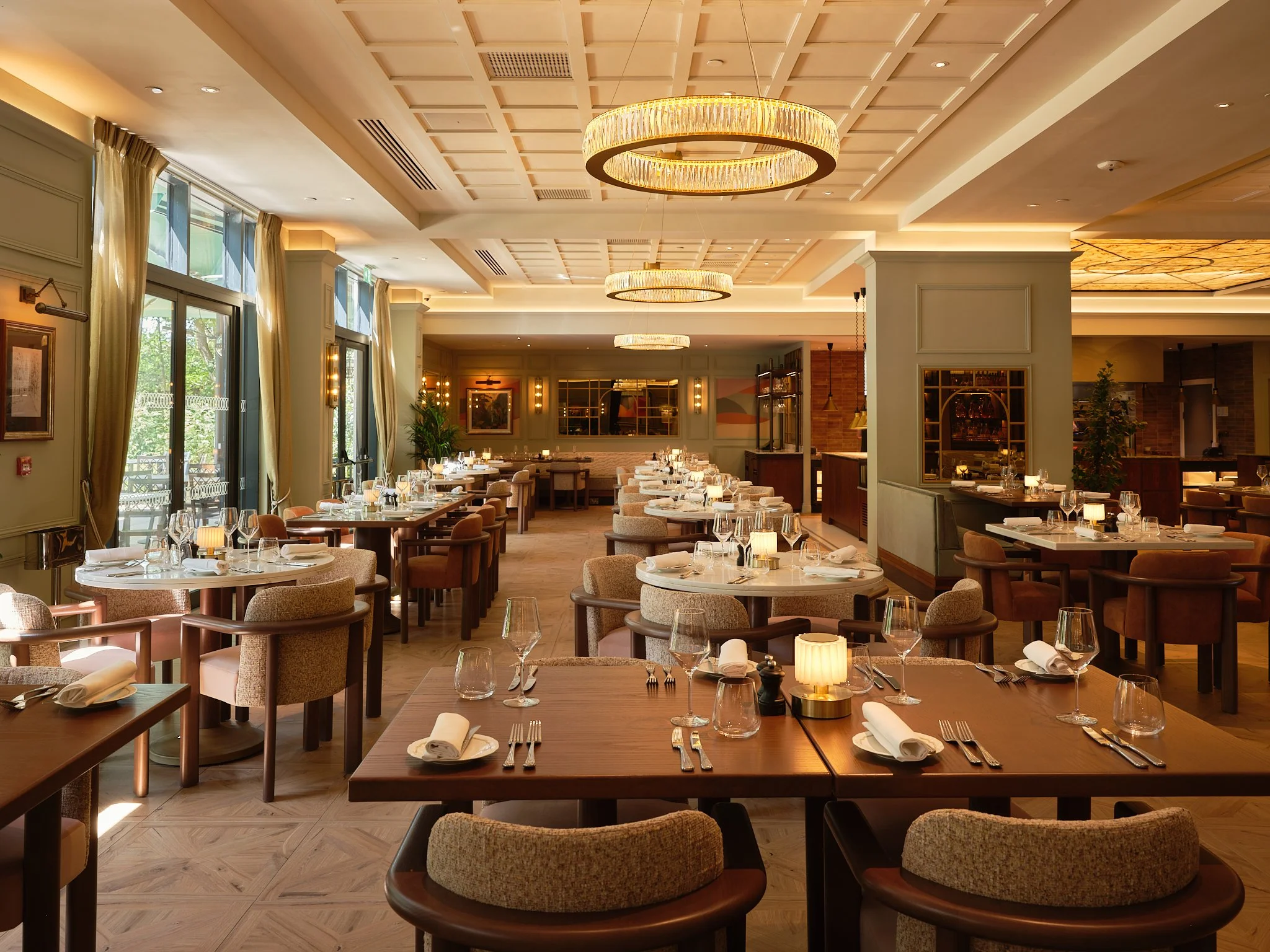 Elegant dining room with round tables set with glassware, silverware, and napkins, surrounded by upholstered chairs. Large windows with curtains provide natural light, and modern circular chandeliers hang from the coffered ceiling. The decor includes framed artwork, greenery, and warm lighting.