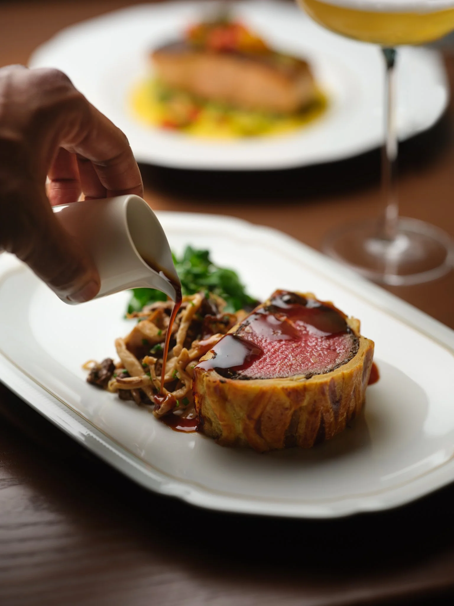 A plate with a slice of beef Wellington surrounded by mashed potatoes and vegetables, garnished with sauce. In the background, a glass of white wine and another plate with a similar dish are visible.