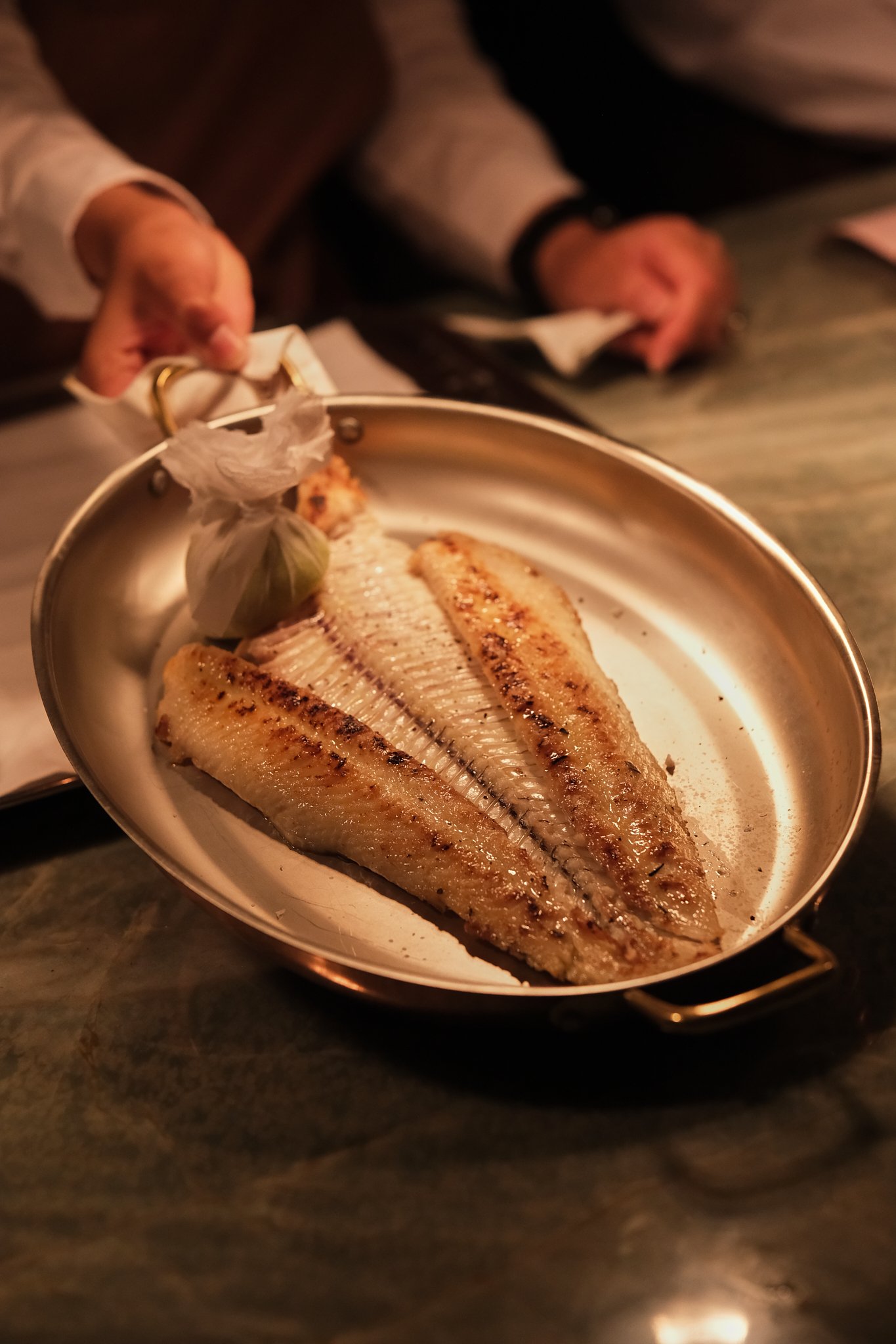 A cooked fish fillet on a metal plate held by a person in a white shirt, with another person in the background.