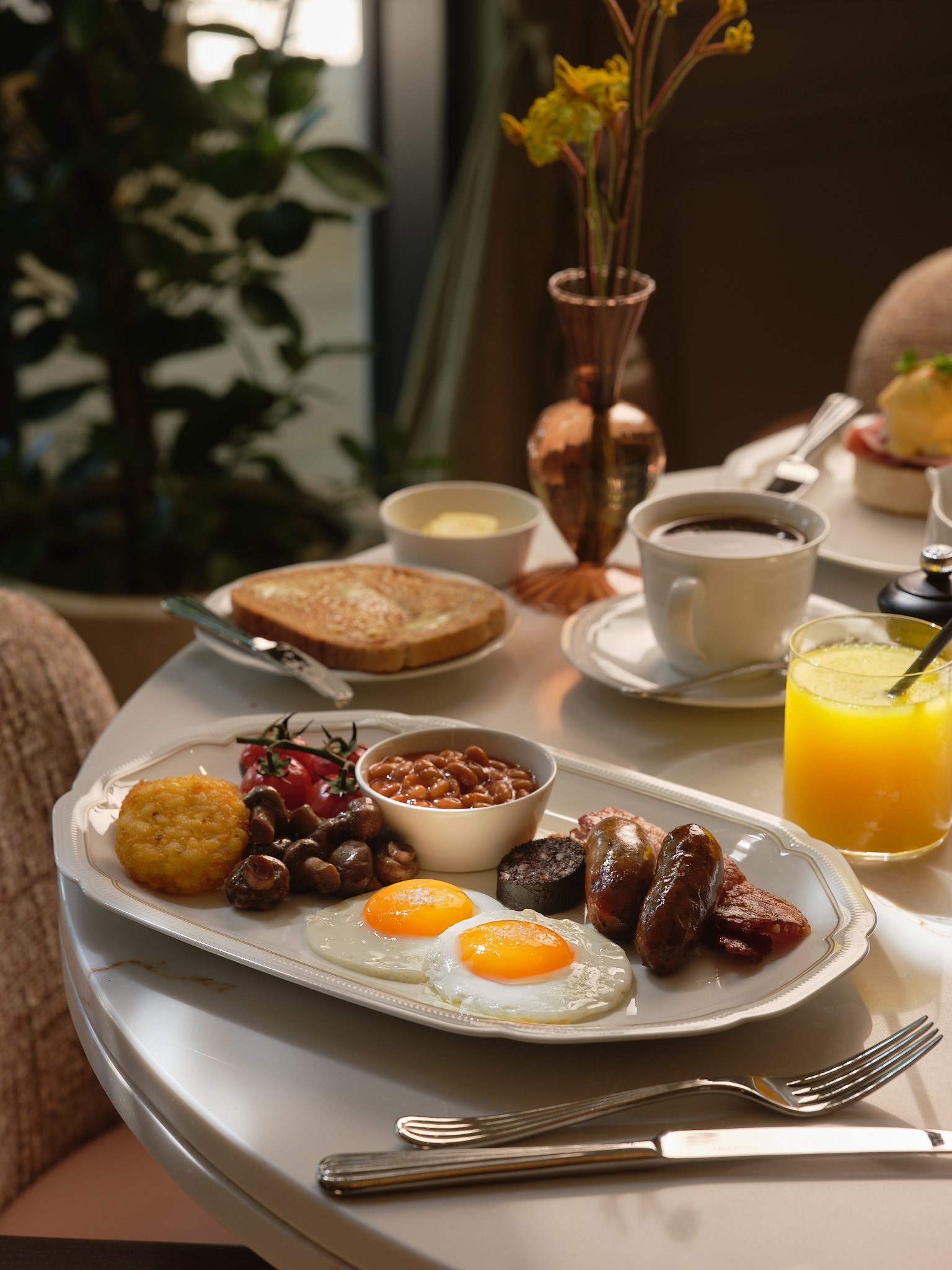 A breakfast meal on a white tray with two fried eggs, sausage links, baked beans, hash browns, cherry tomatoes, and a slice of toast. There is a glass of orange juice and a cup of black coffee on the table, with a background of a tea table set with plates, cups, and a flower vase.
