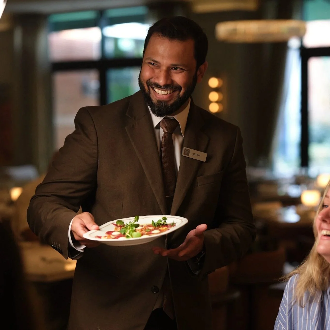 A smiling man in a brown suit and tie with a name tag that says 'Orton' is serving a plate of food to a woman in a restaurant. The background is warmly lit with blurred windows and hanging lights.