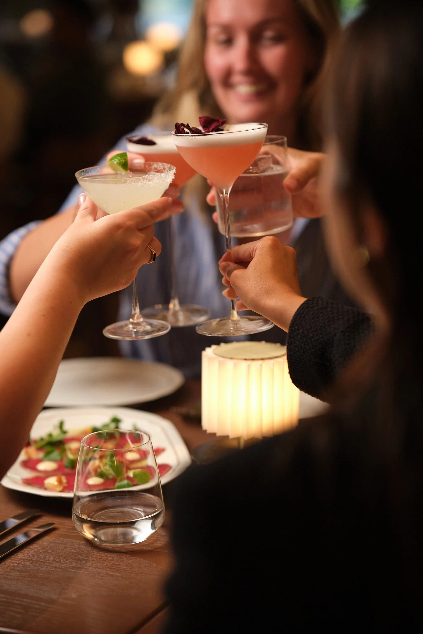 People raising cocktails in a toast at a dinner party, with a plate of food and a glass of water on the table.