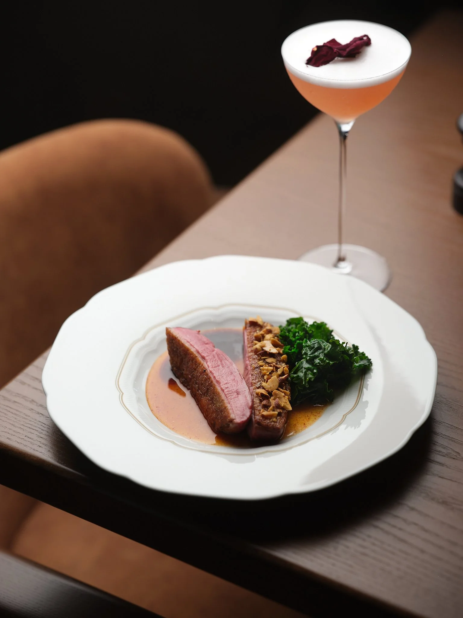 A plated entree with slices of medium-rare beef served with a brown sauce, garnished with chopped nuts, and accompanied by greens; in the background, a cocktail with a frothy top and a garnish of dried fruit.
