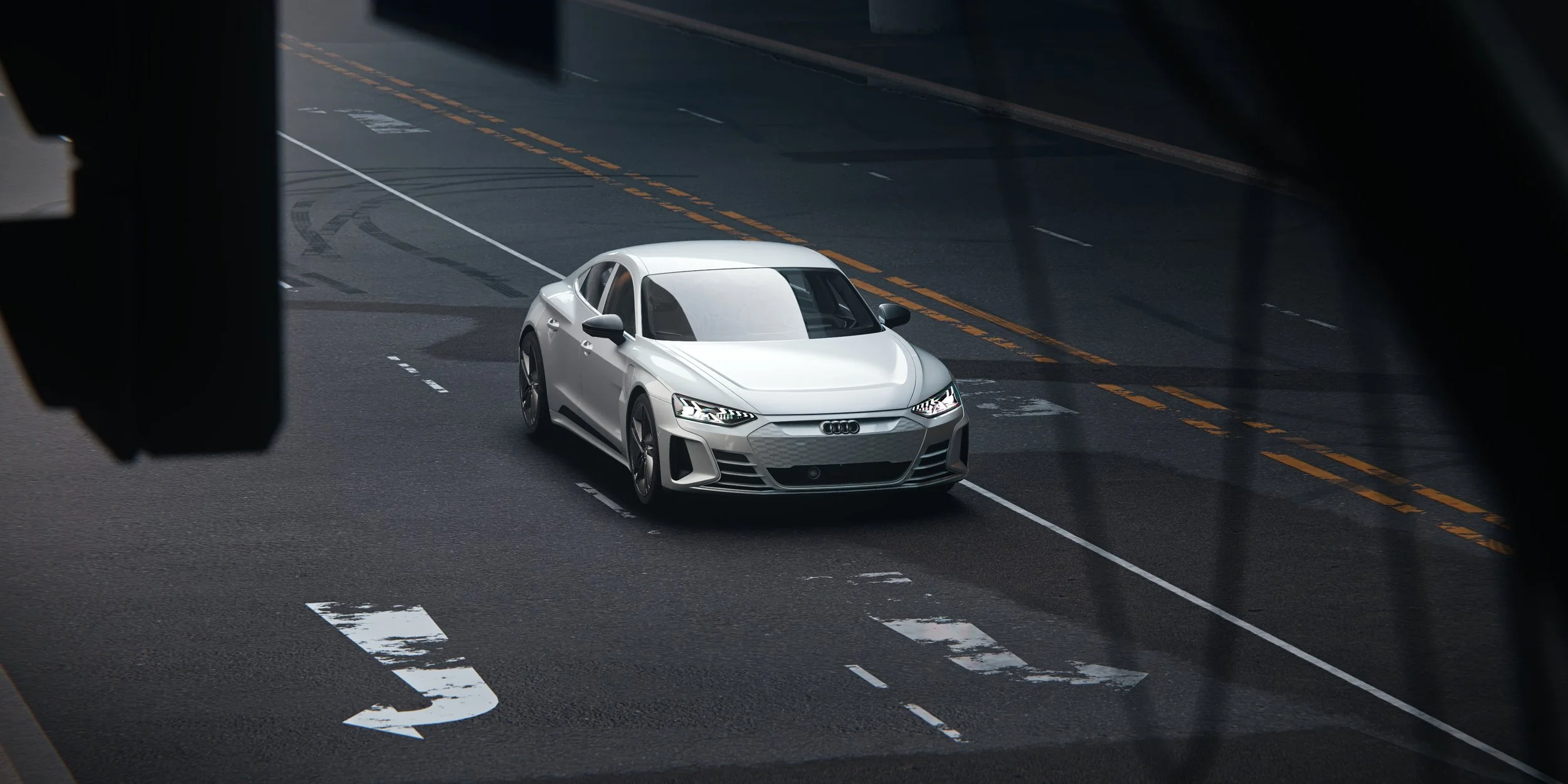 Silberner Audi e-tron GT aus erhöhter Frontperspektive auf einer dunklen Asphaltstraße mit Fahrbahnmarkierungen. Silver Audi e-tron GT viewed from an elevated front angle on a dark asphalt street with road markings.