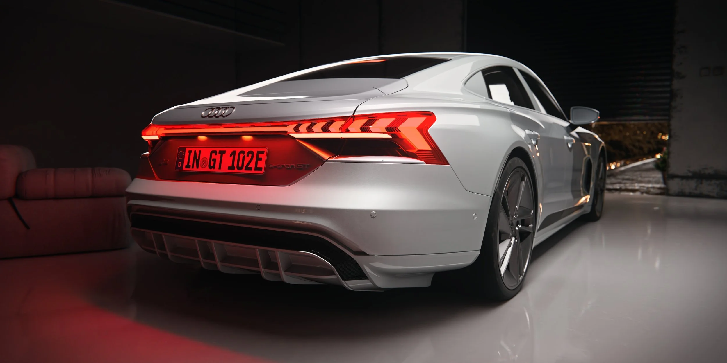 Silberner Audi e-tron GT schräg von hinten mit leuchtendem rotem LED-Lichtband und schwarzem Heckdiffusor. Rear three-quarter view of a silver Audi e-tron GT with an illuminated red LED light bar and black rear diffuser.