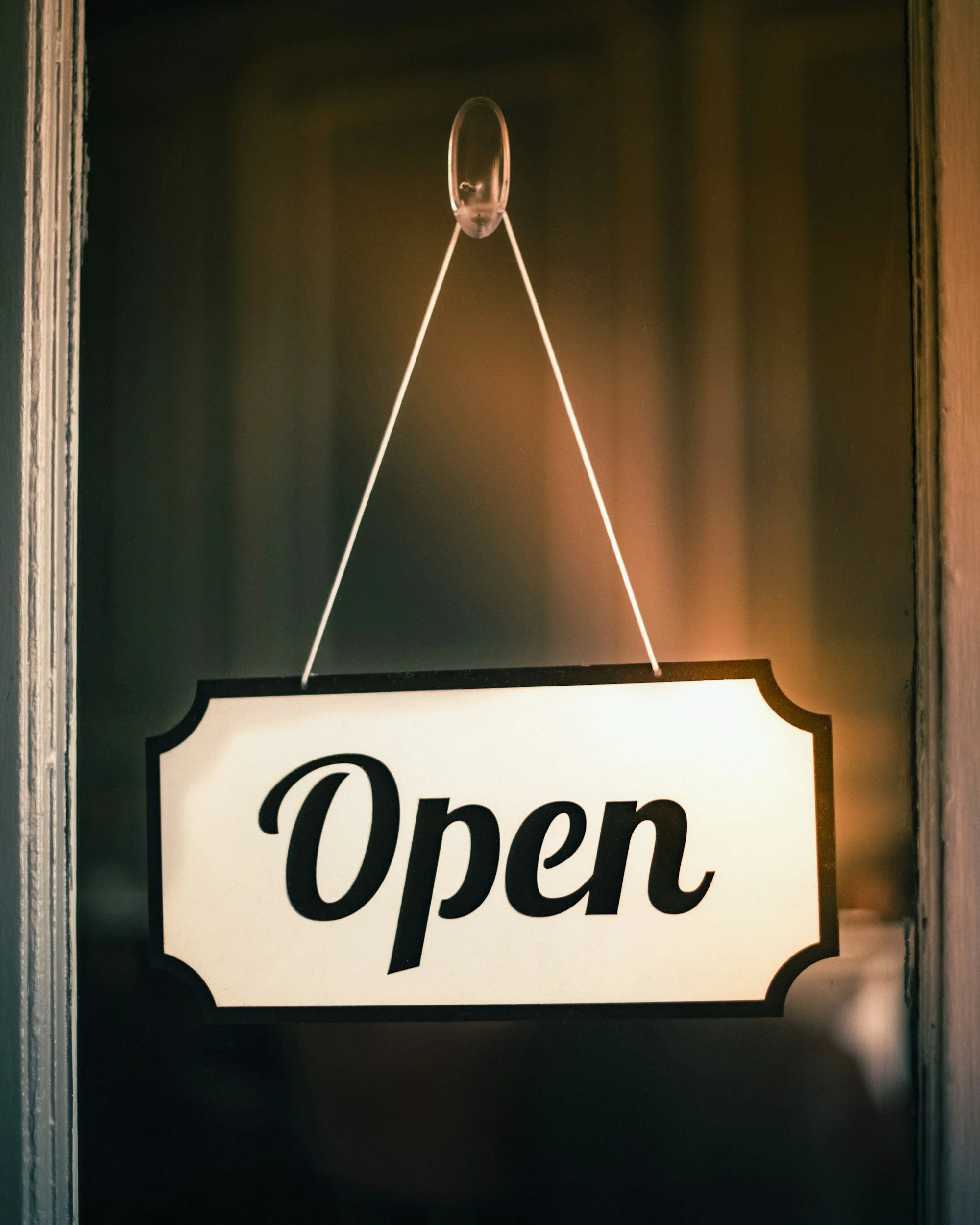 A decorative sign with the word 'Open' in black script on a white background, mounted on a dark wall with a metal frame.