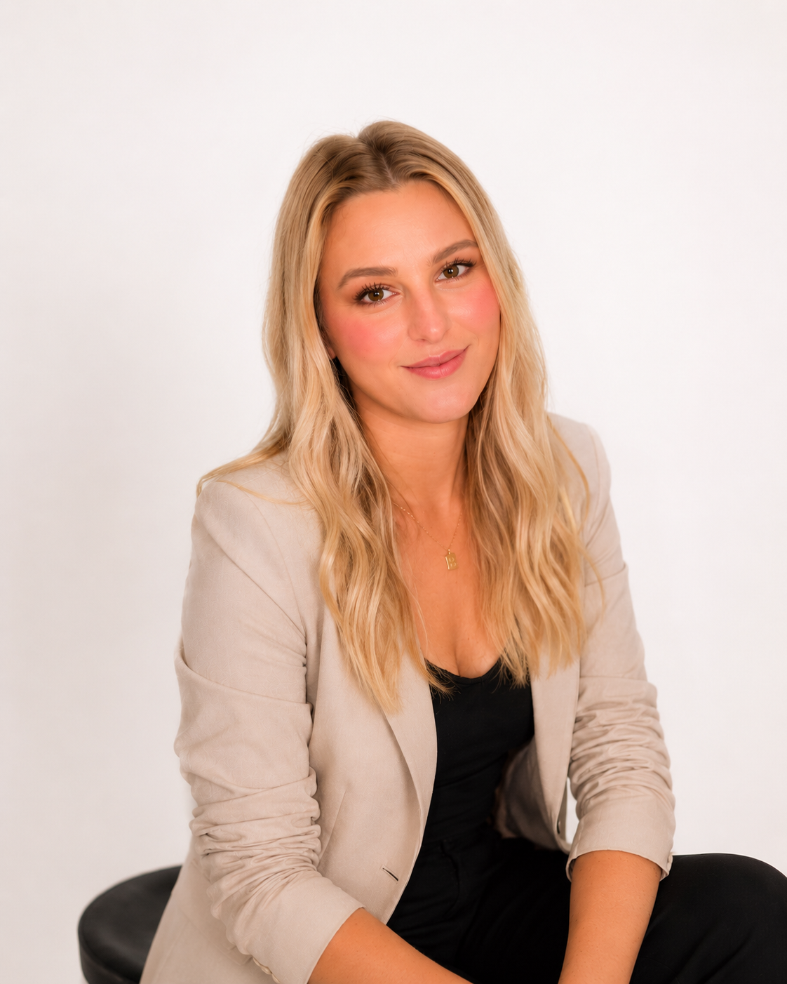 A woman with long blonde wavy hair, wearing a beige blazer over a black top, sitting on a black stool against a plain white background, smiling at the camera.