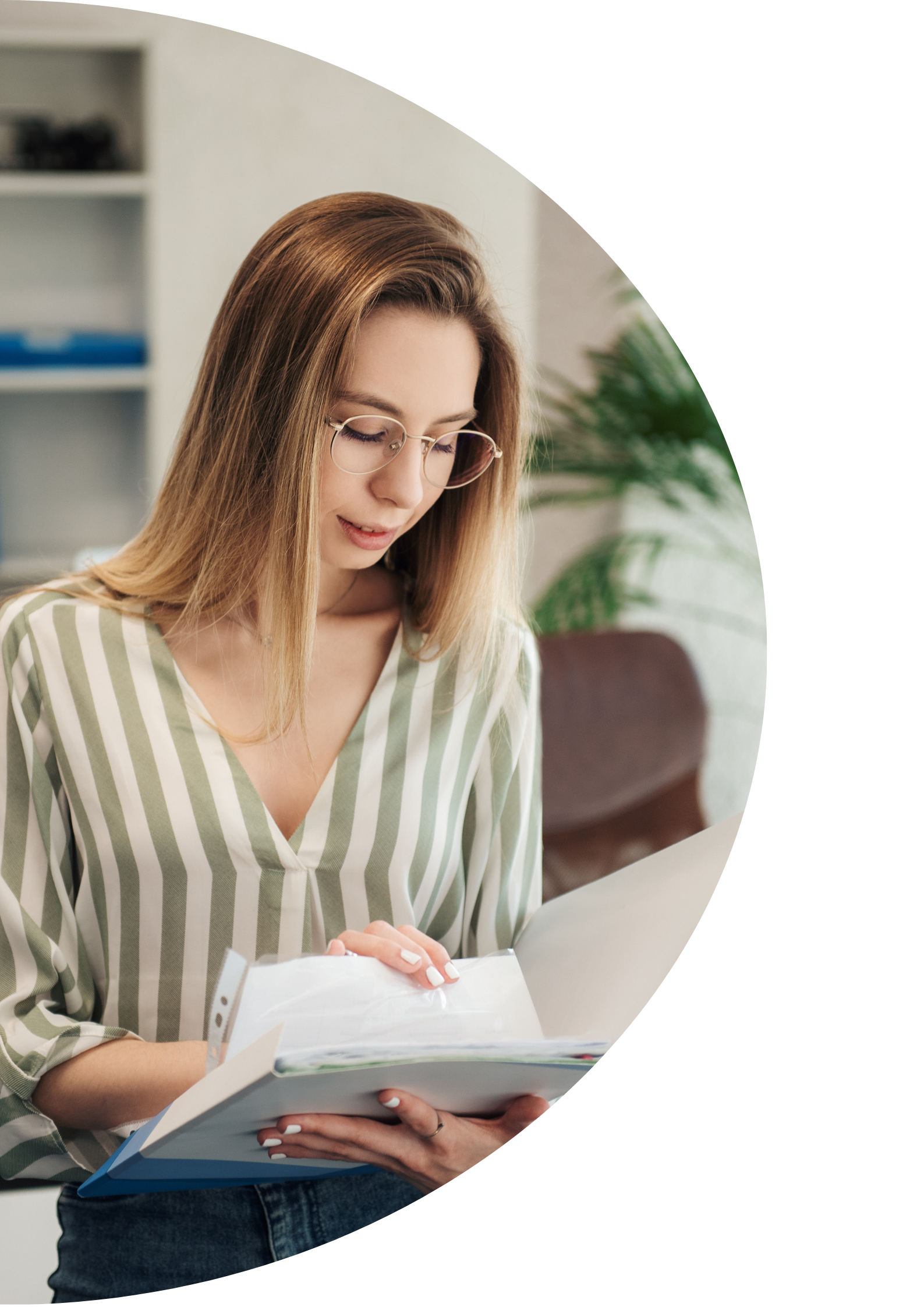 A young woman with long light brown hair wearing glasses and a green and white striped shirt, holding an open book or notebook while standing in an indoor setting with shelves in the background.