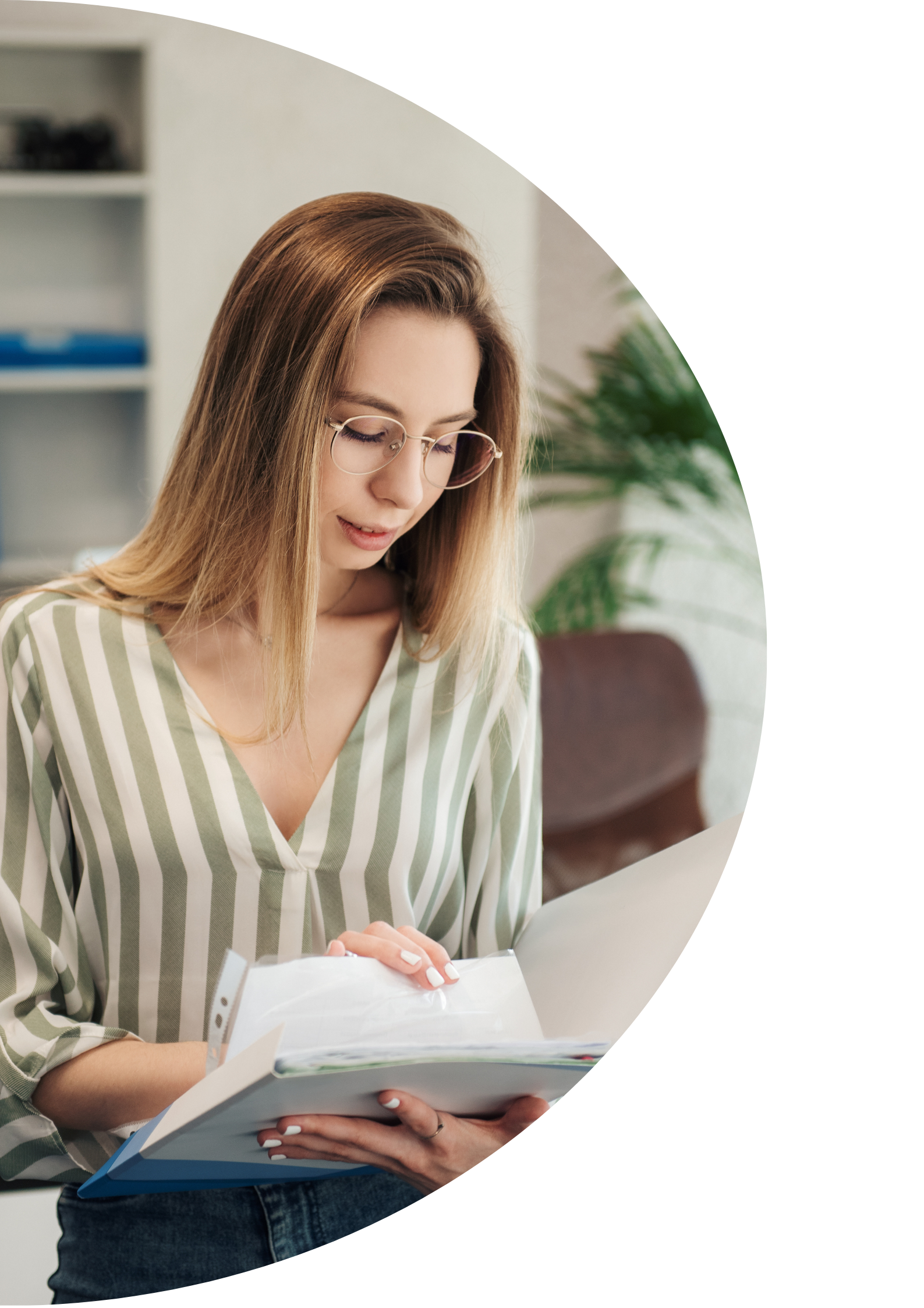 A young woman with light brown hair, glasses, and wearing a striped shirt, is reading a book or notebook in an indoor setting, possibly a library or office.